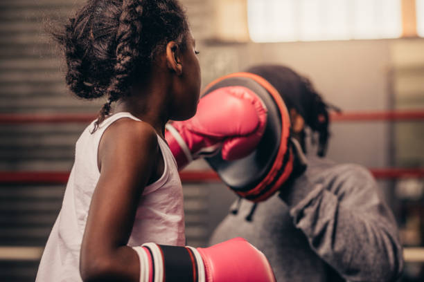 Girl wearing boxing gloves straining with her coach. Boxing kid practicing punches on a punching pad with her coach.